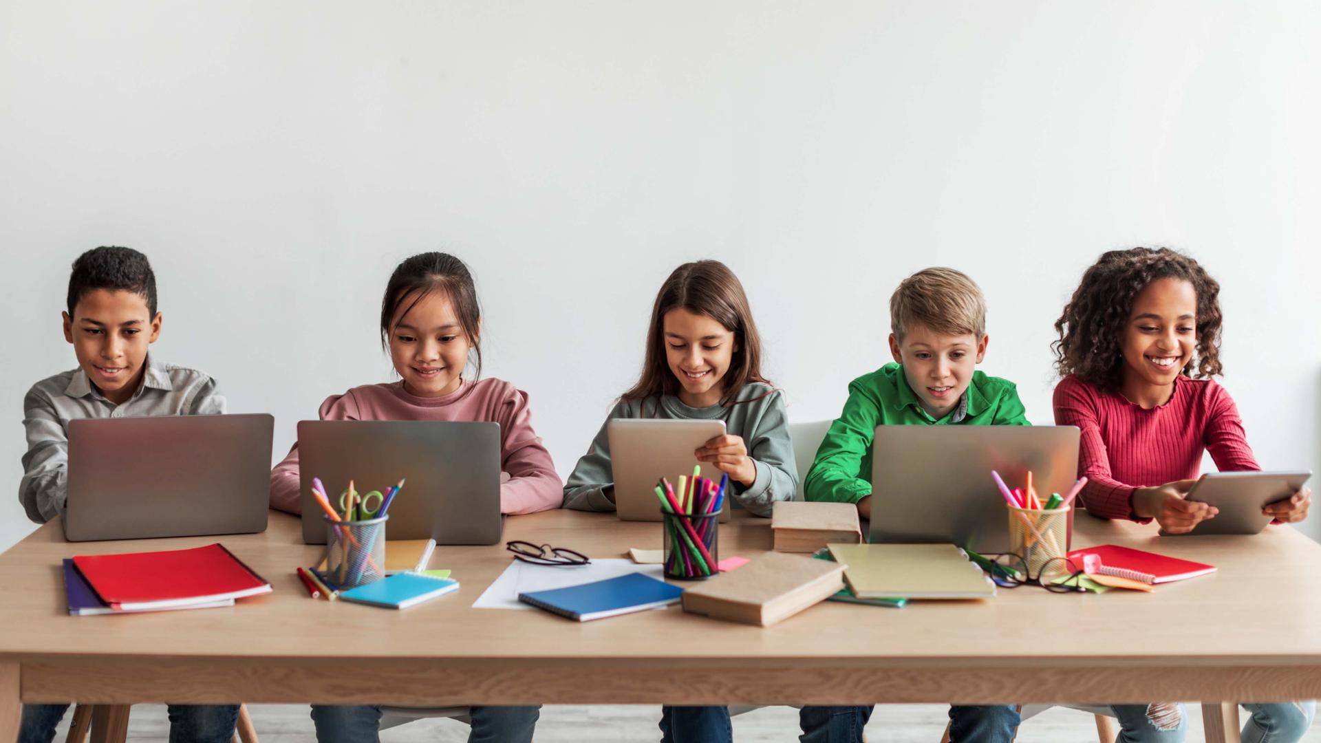 Middle school children sitting at a table, using laptops and tablets.