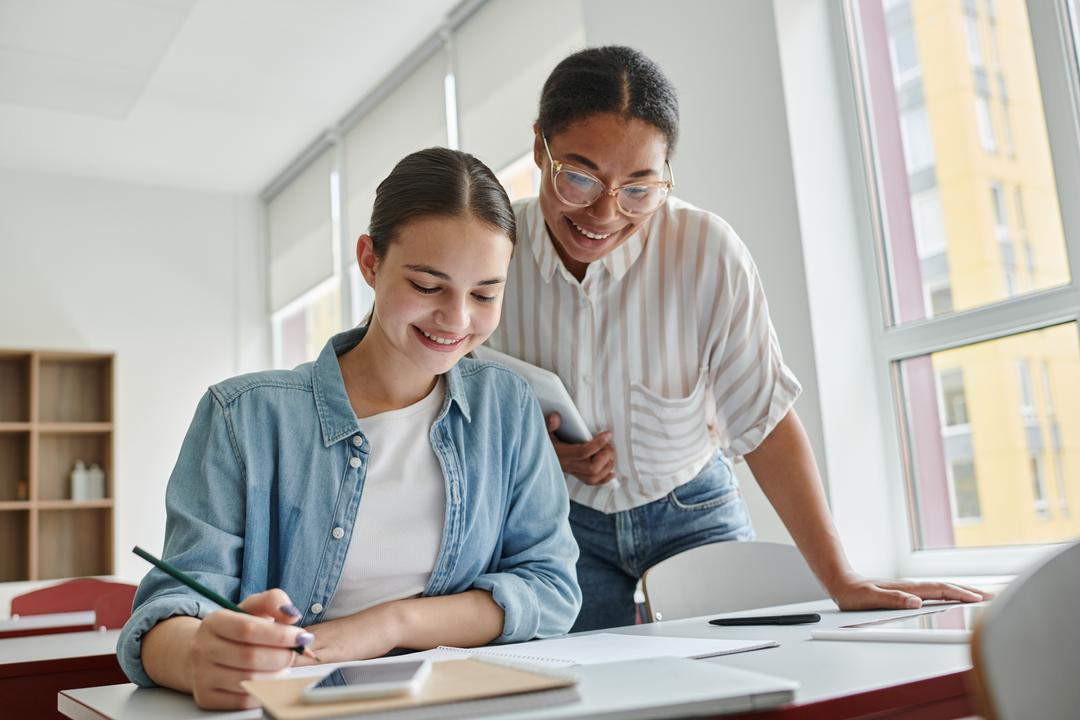 Smiling tutor with digital tablet standing next to teen student doing work on a desk.