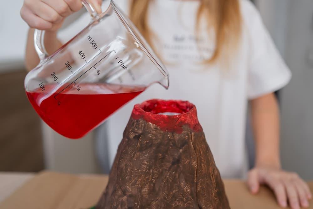 Young girl holding a beaker filled with red liquid above the opening of her volcano science experiment.