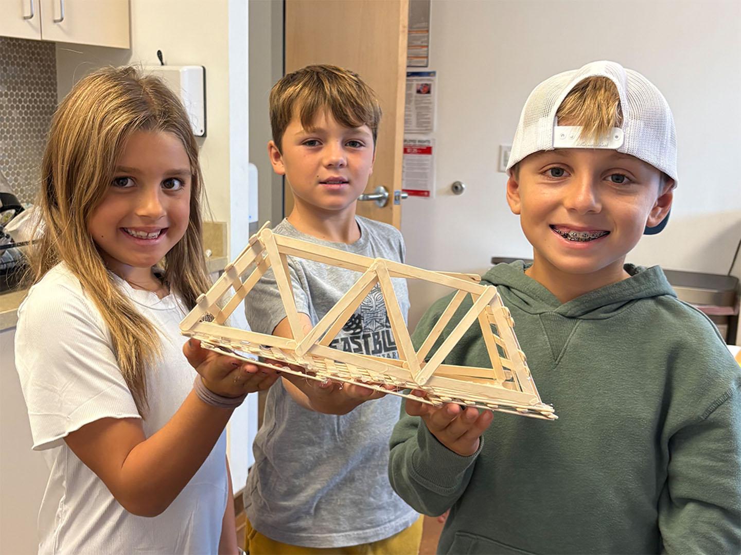 Three children holding a wooden bridge made of popsicle sticks.
